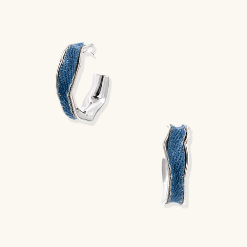 Silver huggie hoops with blue denim fabric inlay strip and polished white gold plated finish on cream background.